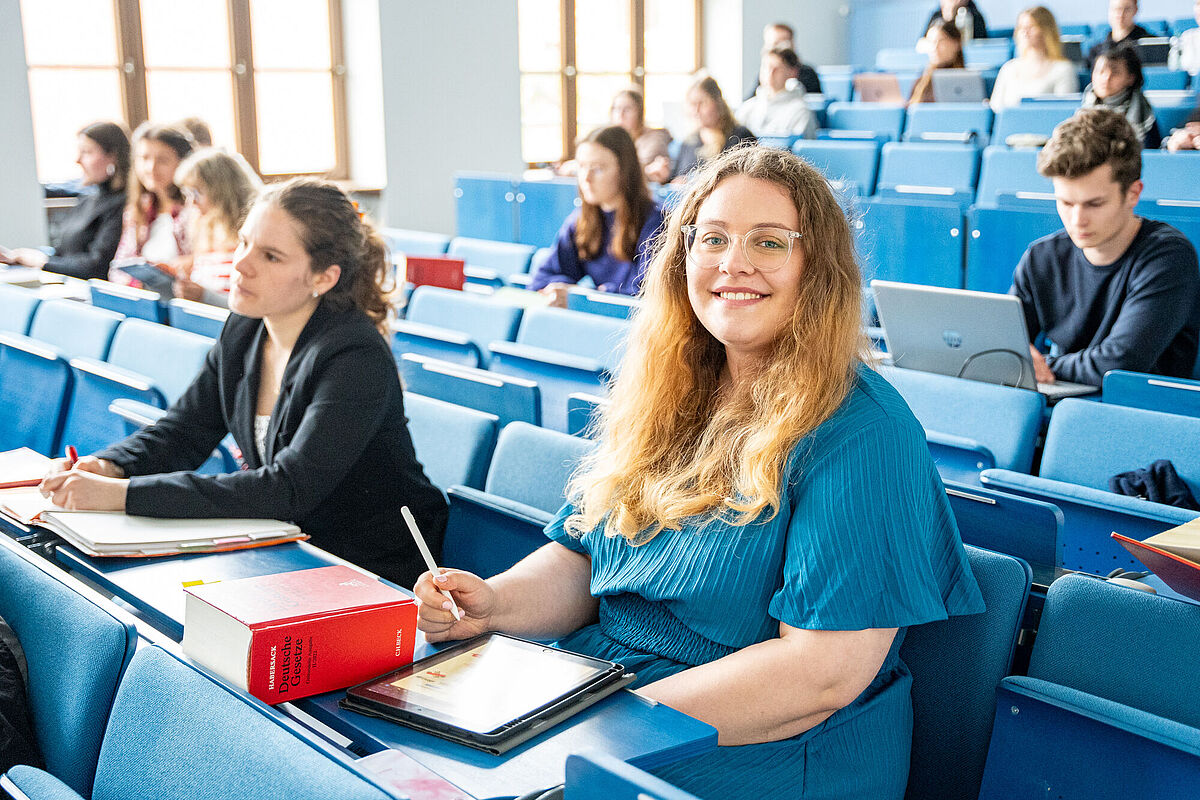 Im Hörsaal sitzen viele Studierende. Im Vordergrund sitzt eine blonde Frau in einem blauen Kleid.